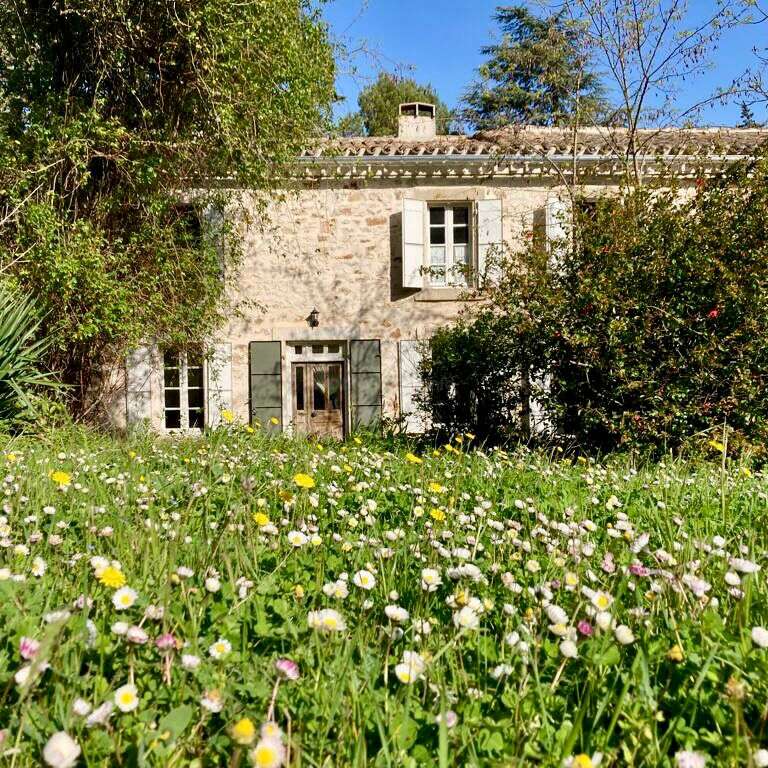 A charming stone house with green shutters sits behind a lush garden filled with wildflowers and tall grass, surrounded by trees under a clear blue sky.