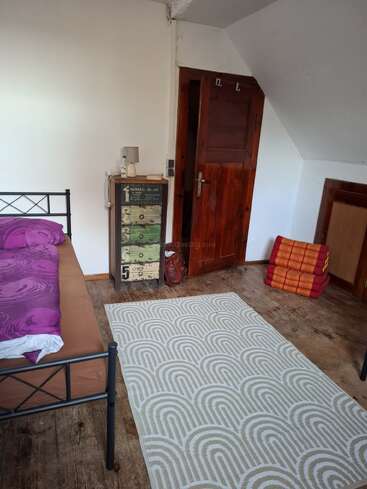 This cozy room features a metal bed with purple bedding, a patterned rug, a colorful chest of drawers, stacked blankets, wooden floor, and a wooden door.