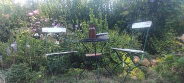 Two white chairs and a small round table sit in a lush, colorful garden. On the table are a brown jug, books, and nature all around.