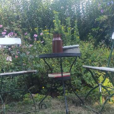 Deux chaises blanches et une petite table ronde sont assises dans un jardin luxuriant et coloré. Sur la table se trouvent une cruche brune, des livres et la nature tout autour.