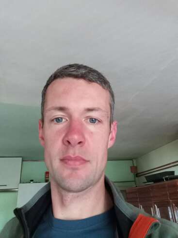 Ein Typ mit kurzen braunen Haaren und blauen Augen ist drinnen und hat eine Jacke an. Im Hintergrund sieht man Küchenschränke, einen Kühlschrank und verschiedene Haushaltsgegenstände.