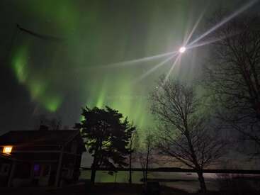 A cozy house glows in the night beneath stunning green northern lights, a bright moon, silhouetted trees, and a calm lake, creating a magical scene.