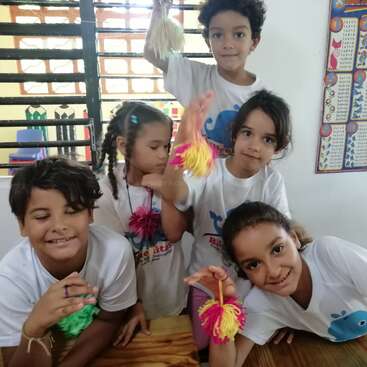 The image shows five children posing for a photo, each holding a colorful yarn pom-pom, in a classroom setting with a window and a calendar on the wall.