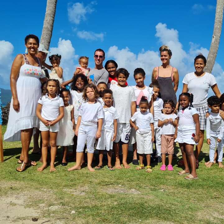The image depicts a group of approximately 20 people, including children and adults, posing for a photo on a grassy area with palm trees and a blue sky with clouds in the background.