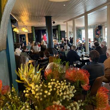 Scène de restaurant animée avec de nombreuses personnes assises à des tables, regardant un artiste sur scène. Éclairage vif, fleurs au premier plan et décor chaleureux.