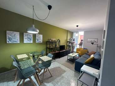 Cette image montre un salon et une salle à manger modernes avec des murs verts, une table en verre, des œuvres d'art, un canapé confortable et deux personnes debout près d'une fenêtre.