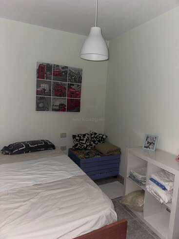 Une petite chambre simple avec un lit simple, des murs blancs, une lampe moderne, des coussins colorés, une étagère avec du linge de maison et une œuvre d'art murale sur le thème de Londres au-dessus du lit.