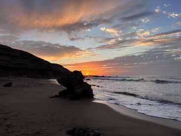 Une scène de plage sereine montre un coucher de soleil spectaculaire, des nuages colorés, de douces vagues s'écrasant sur le rivage, des formations rocheuses et une étendue de sable invitante. Atmosphère paisible.