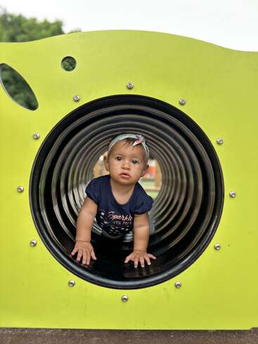 Une petite fille rampe dans un tunnel circulaire de l'aire de jeux, entouré de murs jaune-vert lumineux. Elle porte un bandeau et une chemise bleu marine, et regarde curieusement vers l'avant.