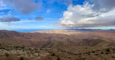 Cette image montre un vaste paysage aride avec des collines rocheuses et une végétation clairsemée sous un ciel spectaculaire, avec de gros nuages et des taches d'horizon bleu clair.