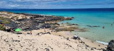 Une belle scène de plage avec une eau turquoise claire, du sable blanc, des roches volcaniques noires, des personnes éparpillées qui se baignent et se détendent, et un parasol vert qui fait de l'ombre sous un ciel lumineux.