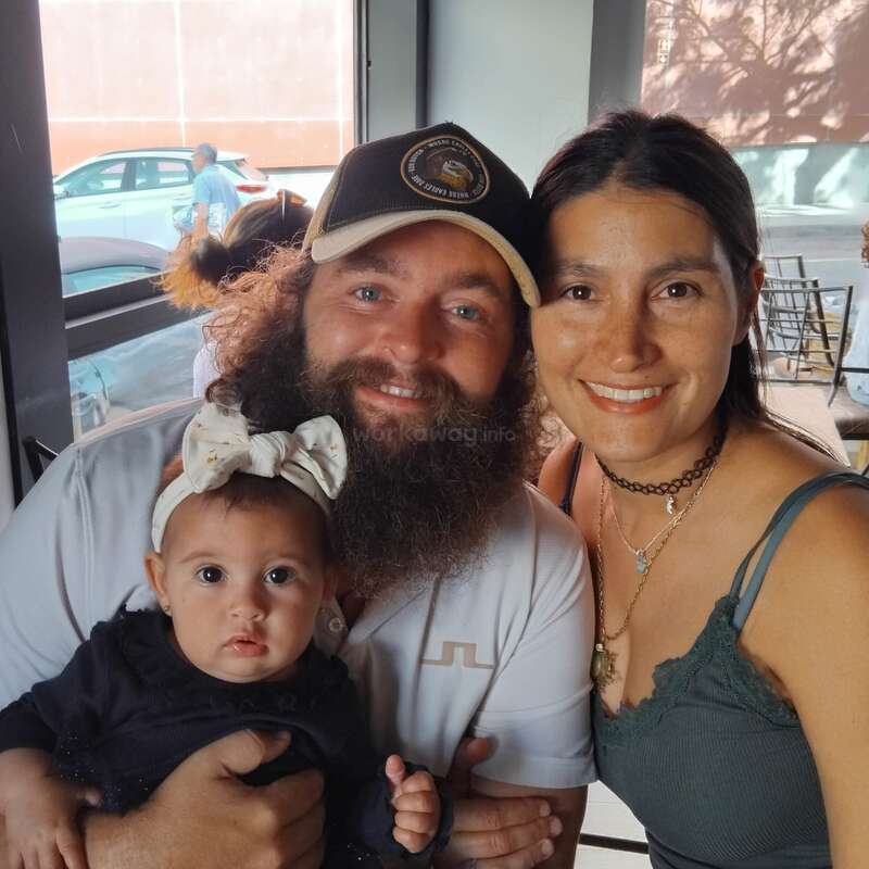 Un homme barbu, une femme et un bébé posent ensemble, souriant chaleureusement à l'intérieur. Le bébé porte un bandeau. La lumière du soleil filtre à travers la fenêtre, créant une atmosphère chaleureuse et joyeuse.