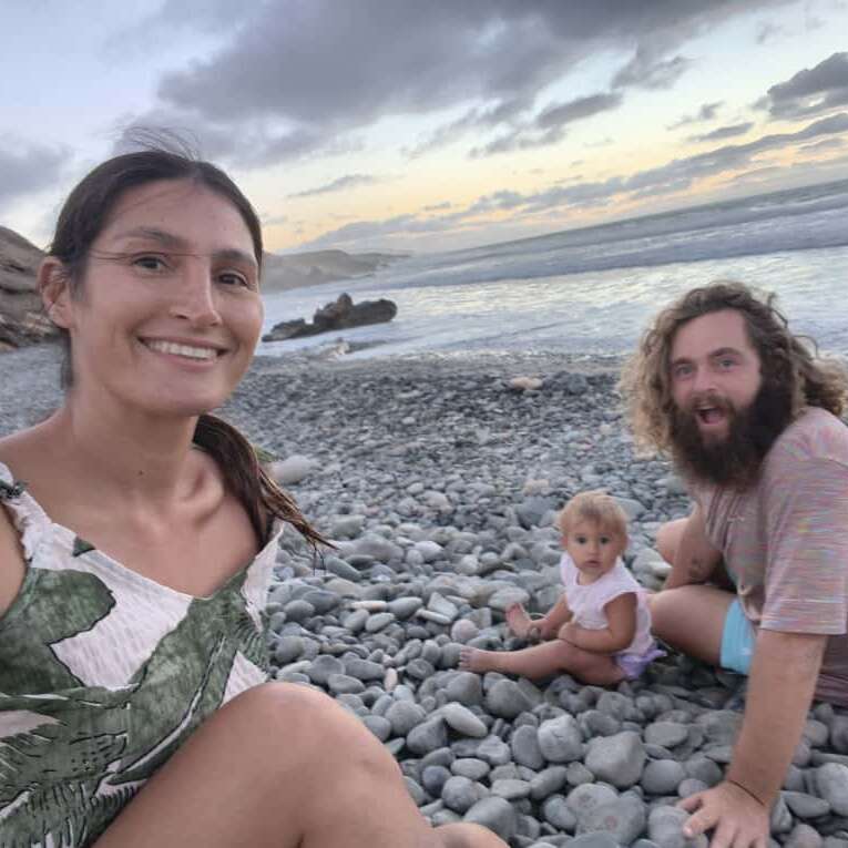A happy family sits together on a rocky beach at sunset. The ocean waves, cloudy sky, and relaxed atmosphere create a peaceful, joyful moment by the sea.