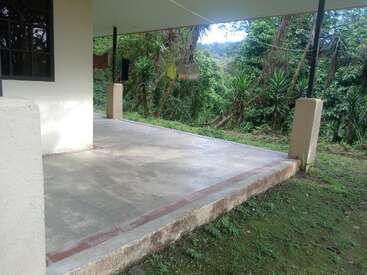Eine Terrasse aus Beton geht von einem Haus in einen üppigen, grünen Wald über. Wäsche hängt auf einer Leine, und Sonnenlicht fällt durch die Bäume und Pflanzen in der Nähe.