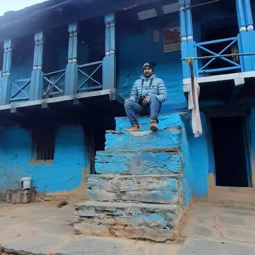 Un hombre sentado en los escalones de piedra de una casa tradicional pintada de azul, con chaqueta y gorro de invierno. El entorno rústico tiene barandillas de madera y telas colgantes.