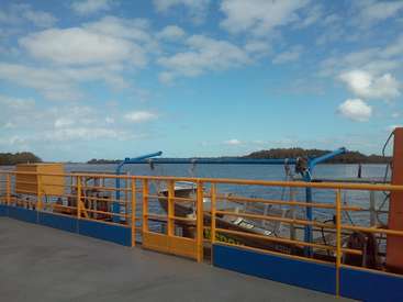 La imagen representa un transbordador con una barandilla amarilla y detalles azules, atracado en un muelle en un día soleado, con una masa de agua y árboles al fondo.