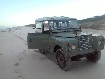 La imagen muestra un Land Rover verde de época aparcado en una playa de arena, con una serena vista del océano y un cielo azul despejado de fondo.