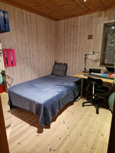 Dieses gemütliche Zimmer aus Holz hat ein Einzelbett mit einer blauen Bettdecke, ein gemustertes Kissen, einen Eckschreibtisch, einen Bürostuhl, Wandregale und ein Fenster.
