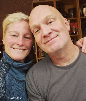 Eine lächelnde Frau und ein Mann machen zusammen ein Selfie in einem Raum. Es ist hell und gemütlich, und im Hintergrund sieht man Regale und verschiedene Sachen.