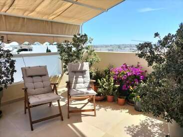 A sunny balcony featuring two cushioned wooden chairs, numerous potted plants, vibrant flowers, and a canopy providing shade, overlooking a cityscape under a clear blue sky.