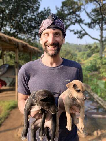 A smiling man wearing a headscarf stands outdoors, holding two adorable puppies, one black and one tan, with greenery and sunlight in the background.