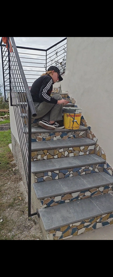 A person sits on outdoor stairs, tiling or grouting the steps with a mosaic of broken tiles. They wear casual clothes and use a yellow bucket.