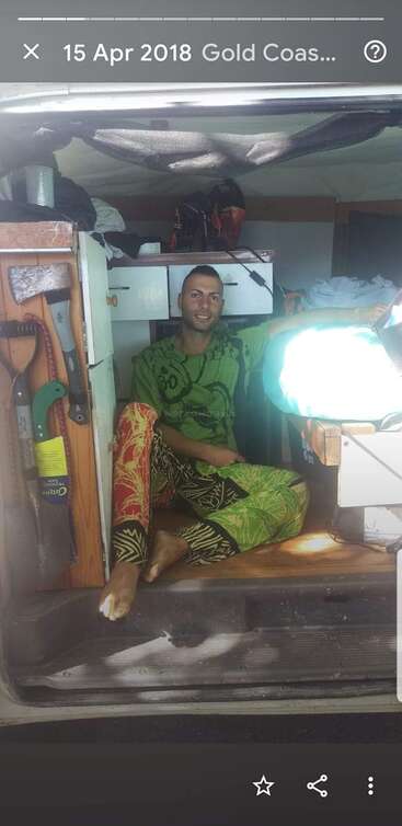 A man wearing colorful clothes sits comfortably inside a van, surrounded by tools, shelves, and bags. Bright sunlight shines in, creating a cozy, relaxed atmosphere.