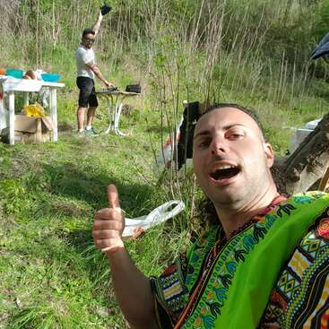 L'image représente deux hommes dans un champ herbeux, l'un cuisinant à une table et l'autre levant le pouce au premier plan.