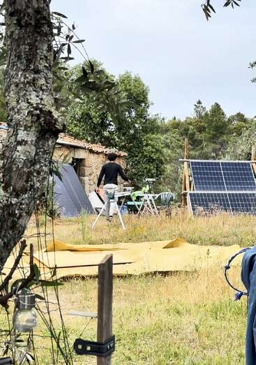 Eine Person arbeitet im Freien in der Nähe von Sonnenkollektoren und einem rustikalen Steingebäude, umgeben von Bäumen und Gras. Auf dem Boden liegen Werkzeuge und gelbe Planen verstreut.