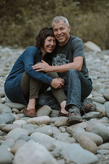Das Bild zeigt ein Paar mittleren Alters, das an einem felsigen Strand sitzt, sich umarmt und in die Kamera lächelt, wobei die Frau ein blaues Hemd und der Mann ein graues T-Shirt trägt.