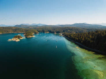 Eine ruhige Küstenbucht mit tiefblauem Wasser, ankernden Booten, bewaldeten Inseln, üppig grüner Uferlinie und fernen Bergen unter einem weiten, klaren, sonnigen Himmel.