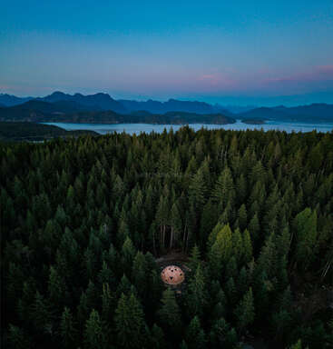 Luftaufnahme einer geodätischen Kuppel inmitten eines dichten Waldes, umgeben von immergrünen Bäumen, mit Bergen und einem ruhigen See in der Ferne.