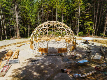 Auf einer Waldlichtung, umgeben von Baumaterialien, Werkzeugen und Arbeitstischen, inmitten von hohen, immergrünen Bäumen, wird eine geodätische Kuppelkonstruktion aus Holz gebaut.