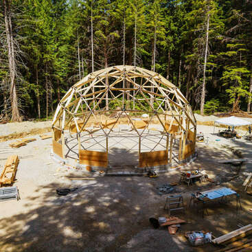 Un dôme géodésique en bois est en cours de construction dans une clairière, entouré de matériaux de construction, d'outils et de tables de travail, au milieu de grands arbres à feuilles persistantes.