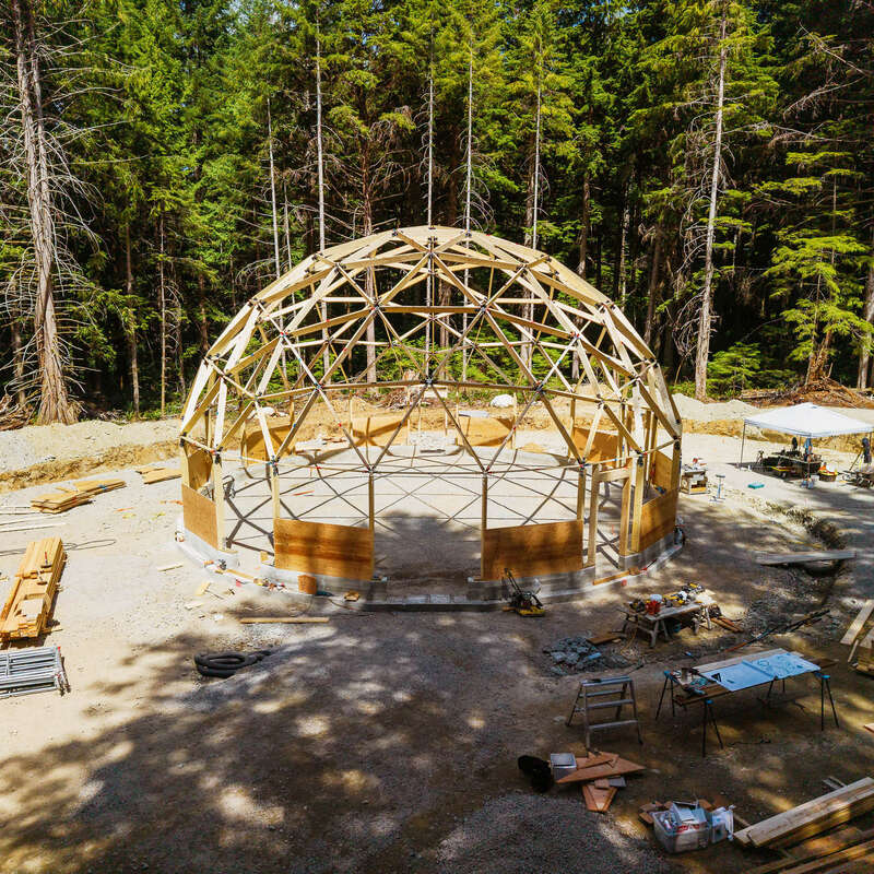 Auf einer Waldlichtung, umgeben von Baumaterialien, Werkzeugen und Arbeitstischen, inmitten von hohen, immergrünen Bäumen, wird eine geodätische Kuppelkonstruktion aus Holz gebaut.
