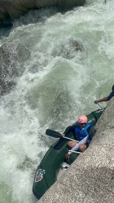 Dos personas con cascos y chalecos salvavidas navegan en una balsa inflable verde por rápidos de aguas bravas, rodeados de grandes rocas y fuertes corrientes. ¡Una aventura emocionante!