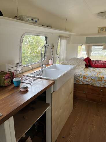 L'image représente l'intérieur d'un camping-car, avec une kitchenette équipée d'un évier, un lit et une fenêtre offrant une vue sur les arbres à l'extérieur.