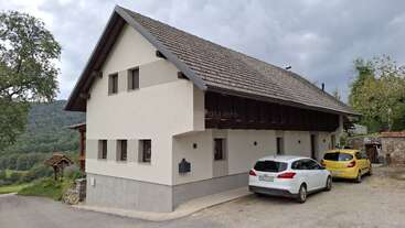 Ein modernes, cremefarbenes Haus mit einem schrägen Dach steht mitten im Grünen. Zwei geparkte Autos - ein weißes und ein gelbes - stehen an einem bewölkten Tag vor dem Eingang.