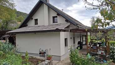 Ein gemütliches, helles Haus mit einem schrägen Dach steht inmitten von üppigem Grün. Im Vordergrund gibt es eine überdachte Veranda mit Gartenmöbeln und einen Gartenbereich.