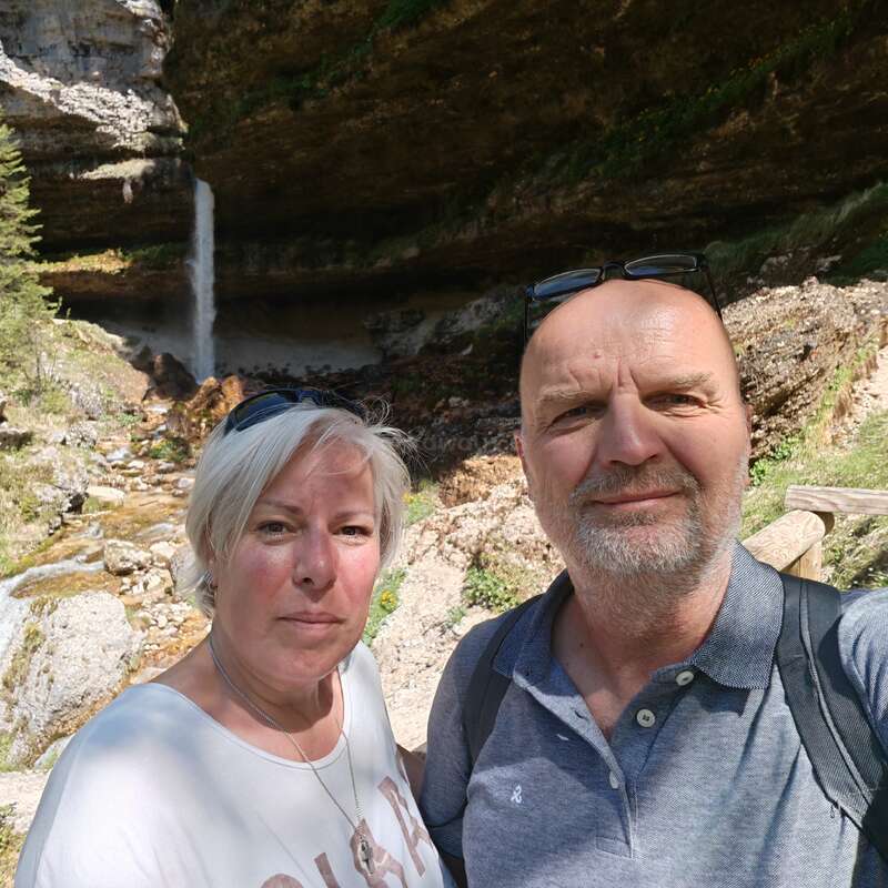 Das Bild zeigt ein Paar mittleren Alters, das für ein Selfie vor einem Wasserfall posiert. Die Frau auf der linken und der Mann auf der rechten Seite tragen beide legere Kleidung.