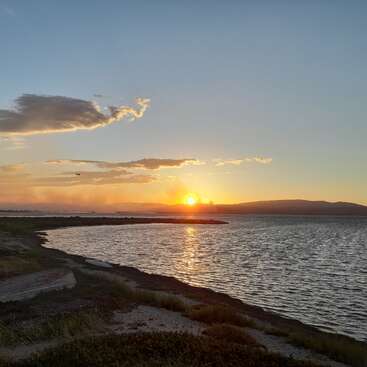 A serene sunset over calm water, with gentle waves, distant mountains, scattered clouds, golden sunlight, a quiet shoreline, and a small airplane in the sky.