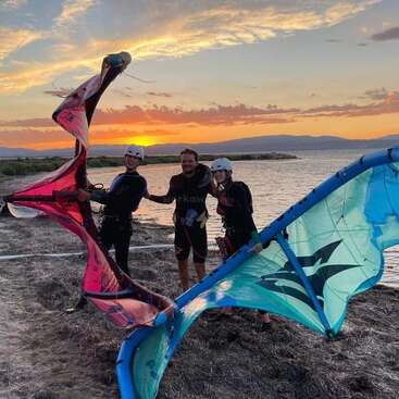 Trois personnes en combinaison de plongée et casques se tiennent sur une plage au coucher du soleil, tenant des voiles de kitesurf colorées, le sourire aux lèvres, avec l'eau et les montagnes en arrière-plan.