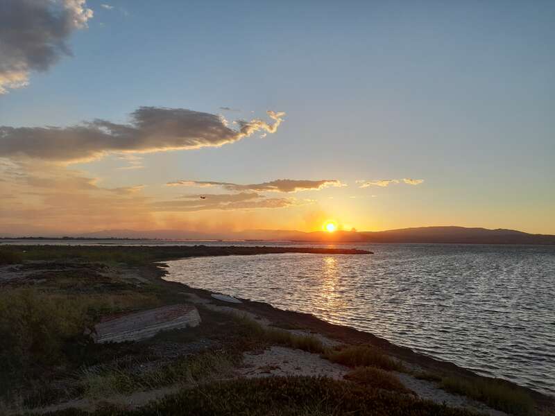 Un coucher de soleil serein sur des eaux calmes, avec des vagues douces, des montagnes lointaines, des nuages épars, un soleil doré, un rivage tranquille et un petit avion dans le ciel.