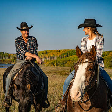 Zwei Leute mit Cowboyhüten reiten auf Pferden über eine Wiese, lächeln sich unter einem strahlend blauen Himmel an und sind von Bäumen in der Ferne umgeben.