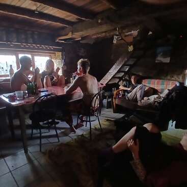 Five people relax in a rustic, dimly lit cabin. Three sit at a table near a window, two lounge on couches, enjoying a cozy, casual atmosphere.