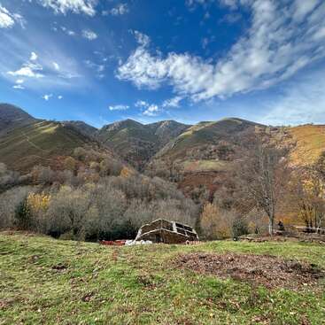 A rustic stone building sits in a grassy field, surrounded by autumn trees, under a dramatic, partly cloudy blue sky, with rolling, green mountains behind.