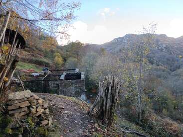 An old stone structure stands amid autumnal trees on a rugged mountain slope. Weathered wooden fence and scattered rocks blend with the tranquil, natural landscape.