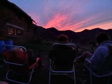 Three people sit in chairs outside a rustic house, watching a spectacular pink and purple sunset over the mountains, enjoying a peaceful, relaxing evening together.