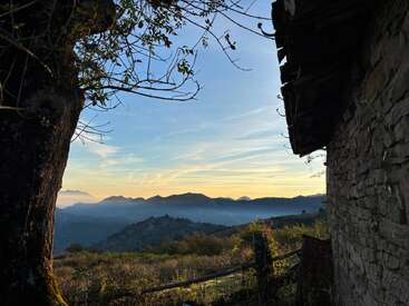 A serene mountain landscape at sunset, with layers of hills, a rustic stone building, a large tree, branches silhouetted, and golden light illuminating the sky.