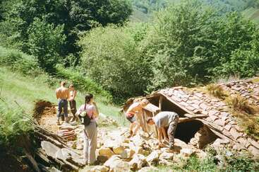 Five people are working together outdoors near a partially collapsed stone building with a tiled roof, surrounded by lush green trees and hills on a sunny day.